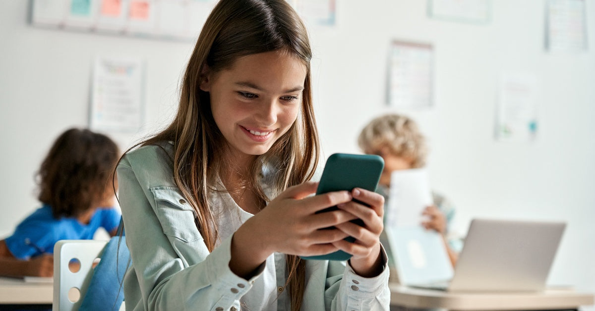 A little girl sits at her desk in a classroom with other children behind her. She smiles as she holds a smartphone.