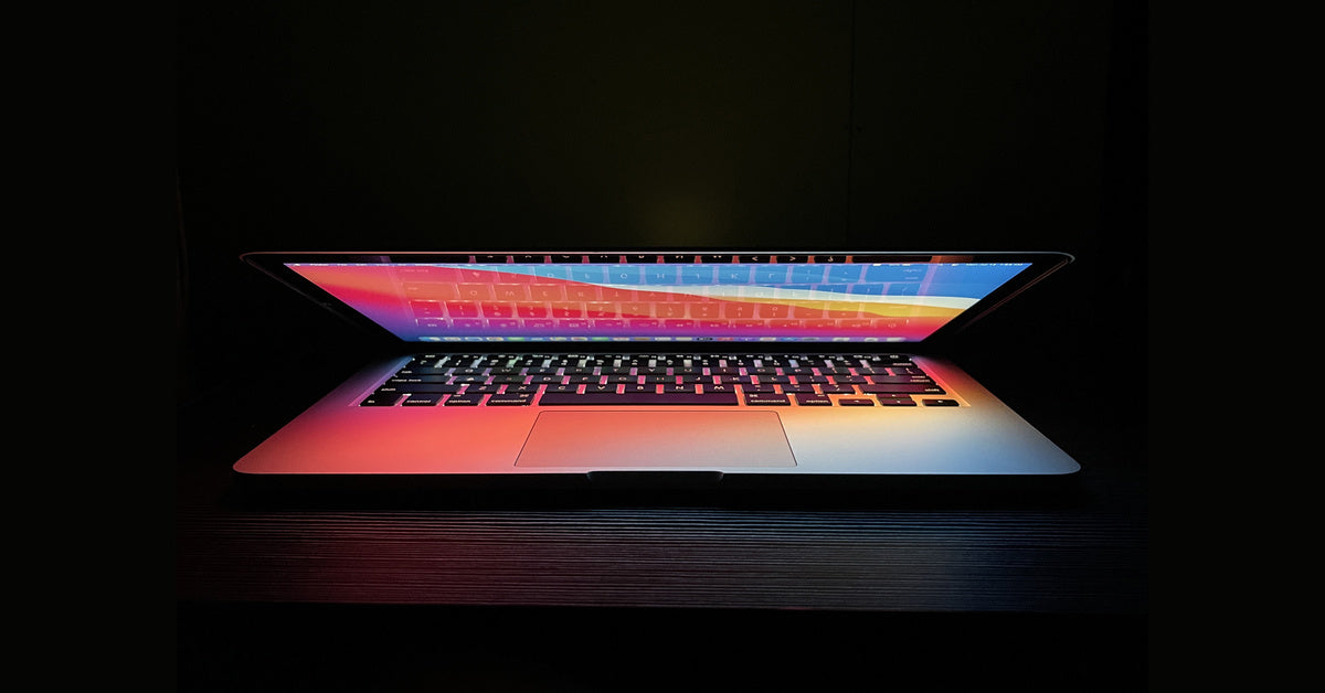 A half-open laptop in a dark room. The display is lit up, casting various colors over the laptop's keyboard.