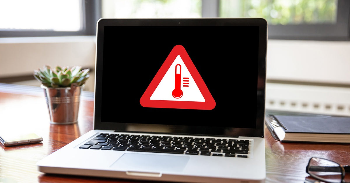 A laptop on a wooden desk with a pen, glasses, a plant, and a notebook nearby. The screen displays a high-temperature warning symbol.